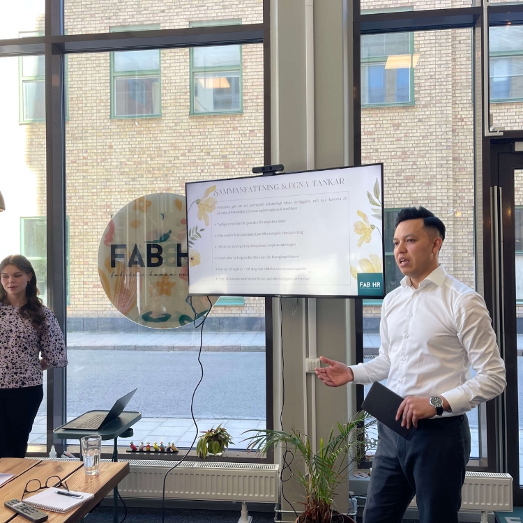 Two people in an office, one presenting in front of a screen with a laptop on the table